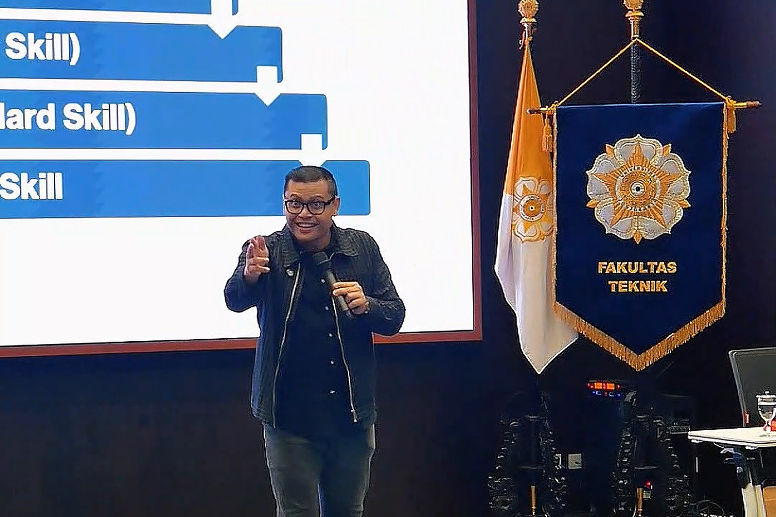 Saya menunjuk ke arah peserta sambil memegang mikrofon di depan layar presentasi dan panji Fakultas Teknik UGM.